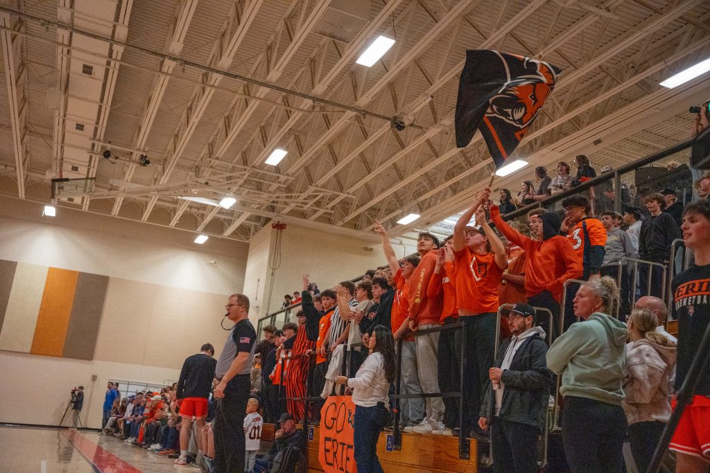 A cheering crowd in orange shirts waves a flag and holds a sign at a basketball game in a gymnasium.