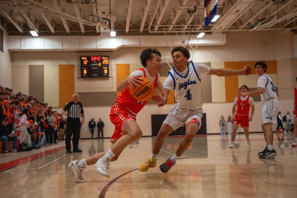 Two basketball players compete for the ball during a game, with a crowd watching in the background. Scoreboard shows 12-20.