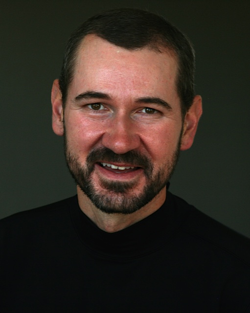 A man with a short beard and short hair smiles while wearing a black shirt against a dark background.