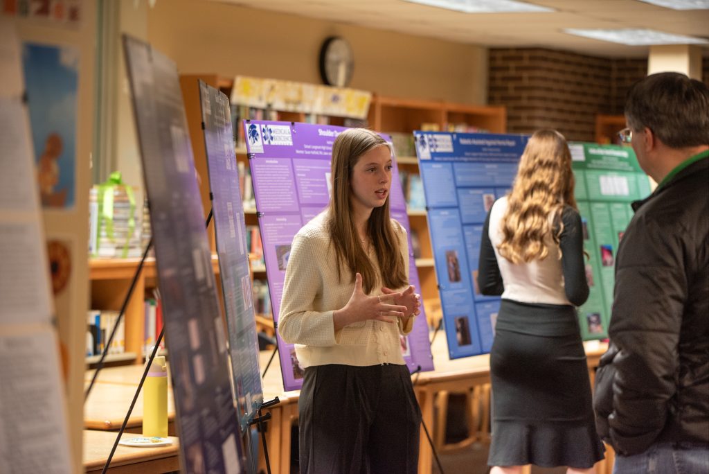 A student presents her project at the MBSA Showcase, with informational posters displayed in a library setting.