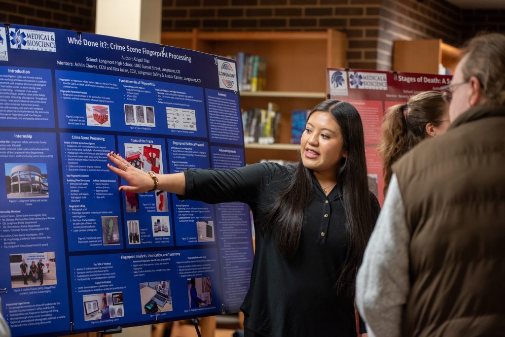 A student presents a research poster on crime scene fingerprint processing to an audience in a library setting.
