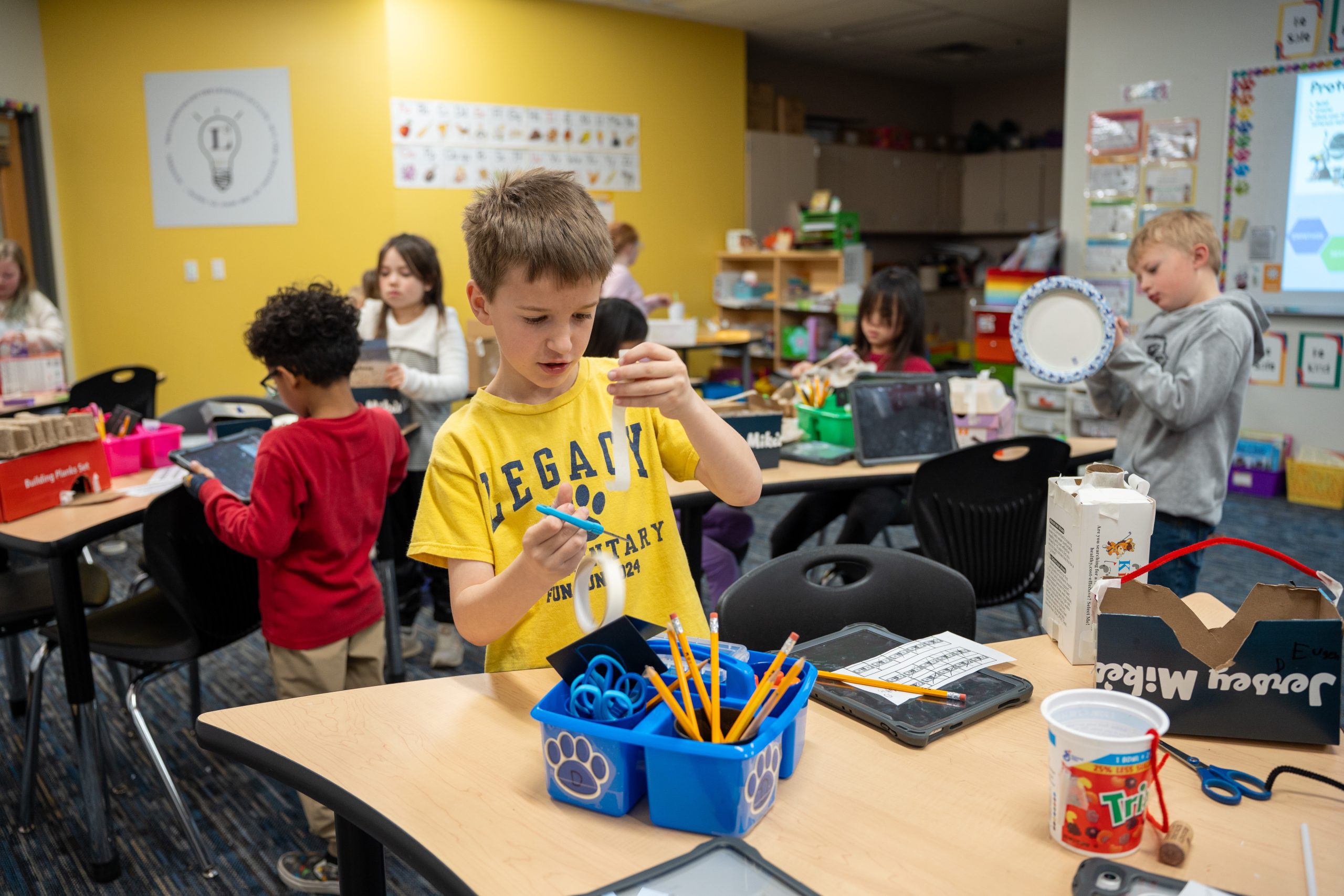 Students are engaged in creative activities at a classroom table, using various art supplies and tools.