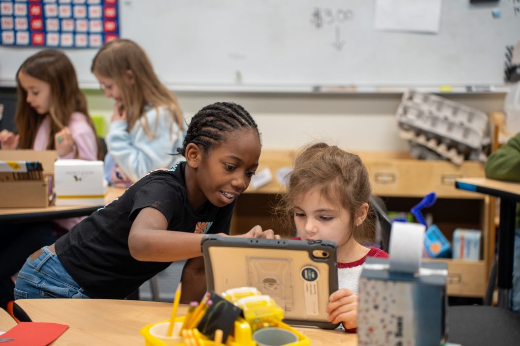 Two children interact with a tablet in a classroom, while other students work at nearby desks.