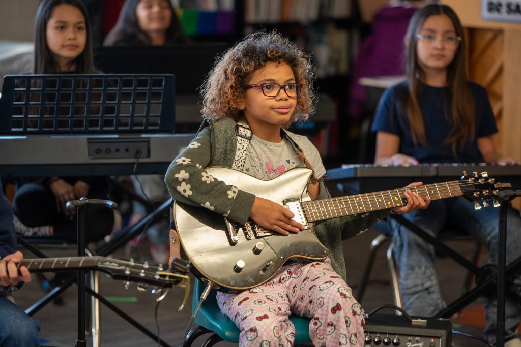 A girl with curly hair and glasses plays a silver guitar, seated among other children in a music class.