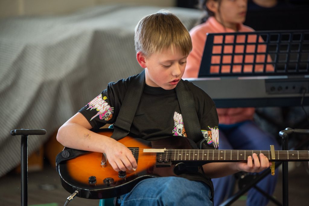 A young boy plays an electric guitar, focused and seated, with peers in the background. He's wearing a black graphic t-shirt.