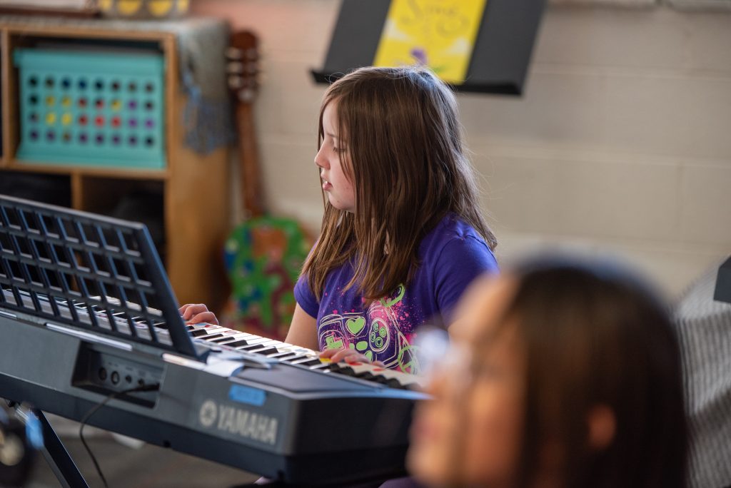 A girl plays a keyboard, focused, in a music class with colorful decor in the background.