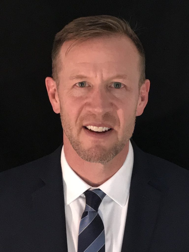 A man with short hair smiles, wearing a dark suit and striped tie, against a black background.