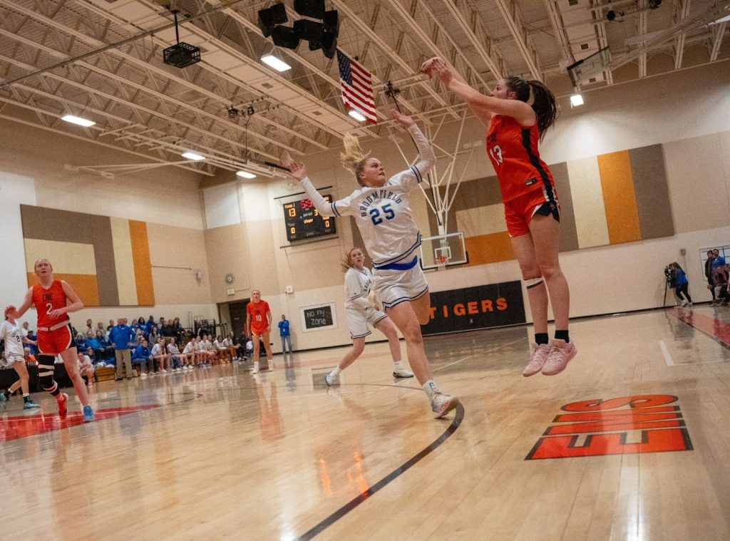 A basketball player in orange jumps to shoot while a defender in white tries to block her. Spectators watch in the background.