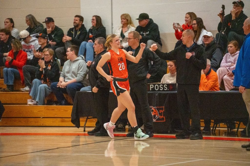 A basketball player in an orange jersey celebrates while coaches and fans cheer in a crowded gym.