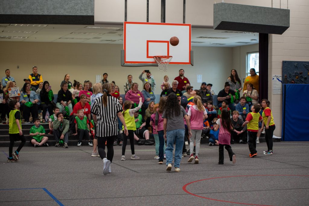 A youth basketball game in progress with players in colorful jerseys and a cheering crowd in the background.
