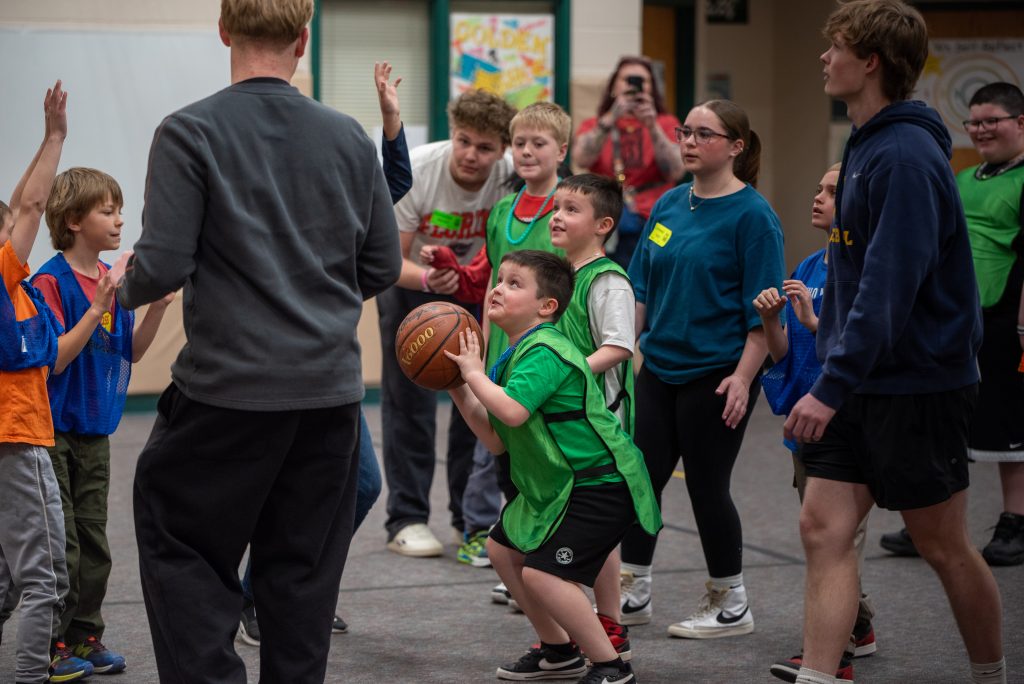 A group of children in a gym, with one boy in green preparing to shoot a basketball while others watch excitedly.
