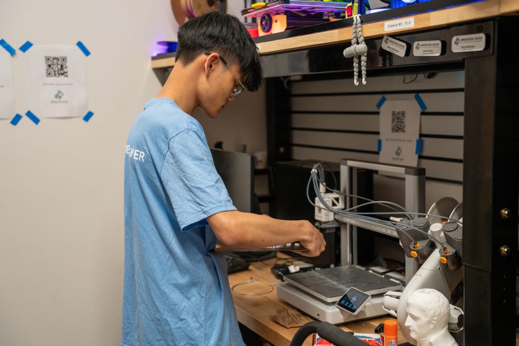 A person in a blue shirt works on a 3D printer in a workshop filled with tools and equipment.