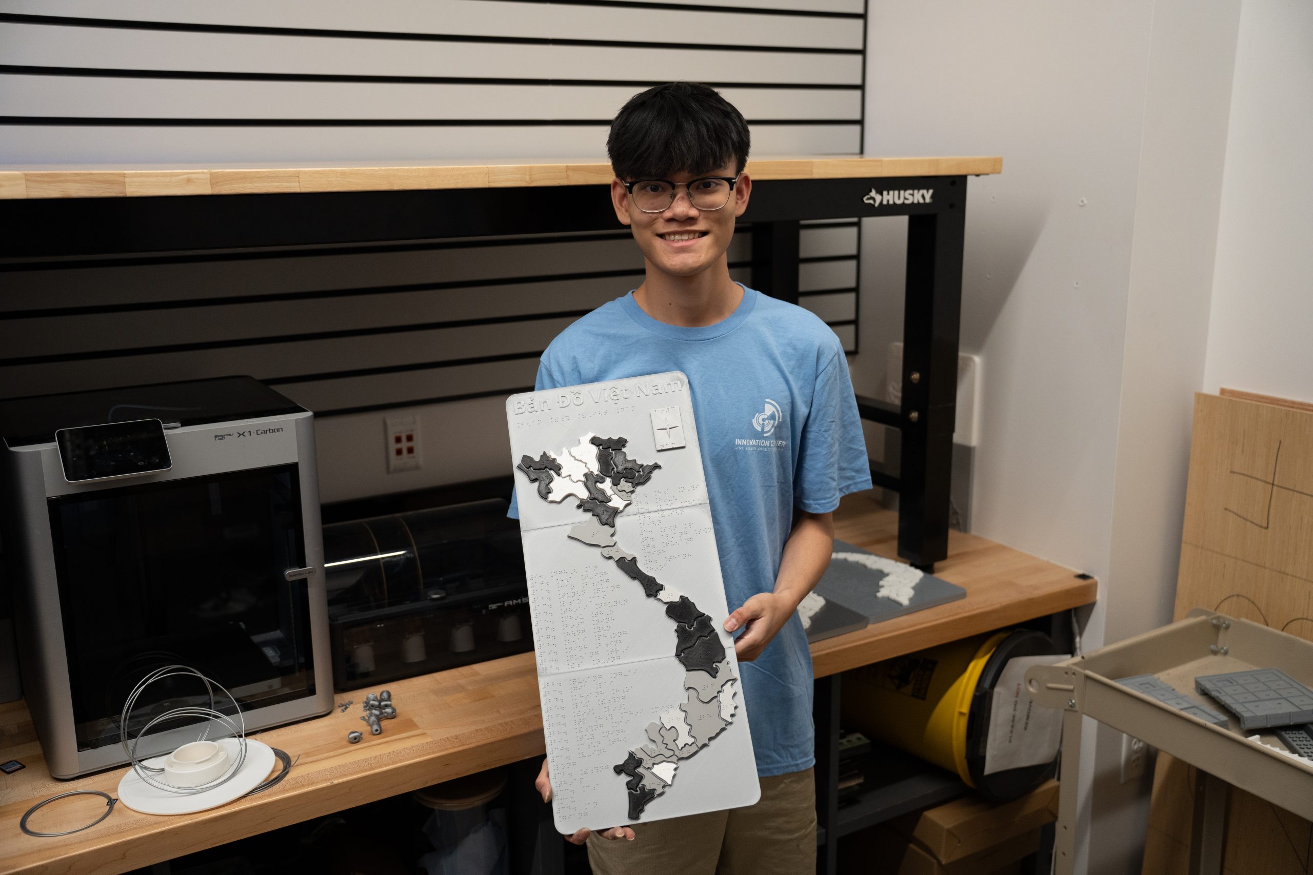 A young man in a blue shirt holds a detailed map of Vietnam in a workshop with tools and equipment in the background.