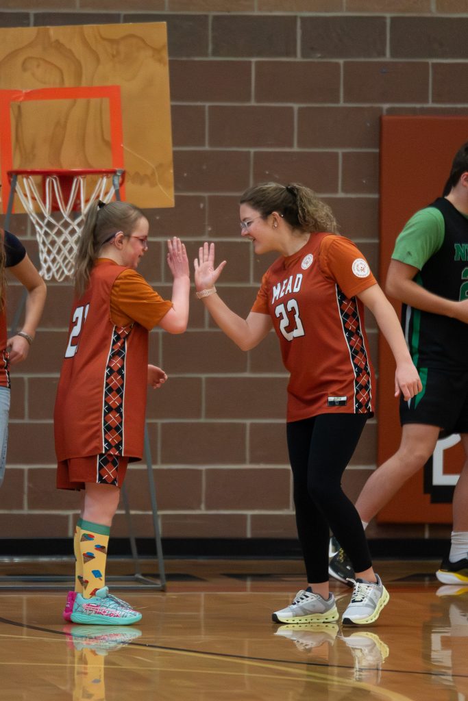 Two girls in basketball jerseys are high-fiving on a court, with a hoop in the background and other players nearby.