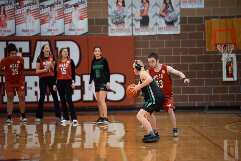 A basketball game scene with players in action, one dribbling while others watch from the sidelines.