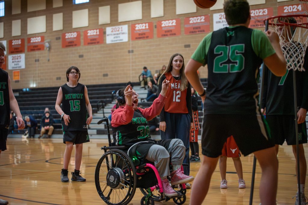 A girl in a wheelchair plays basketball, surrounded by teammates in jerseys, as a ball is shot towards the hoop.
