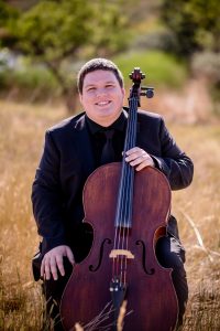 A smiling musician in a black suit sits outdoors, holding a cello amidst tall grass and greenery.