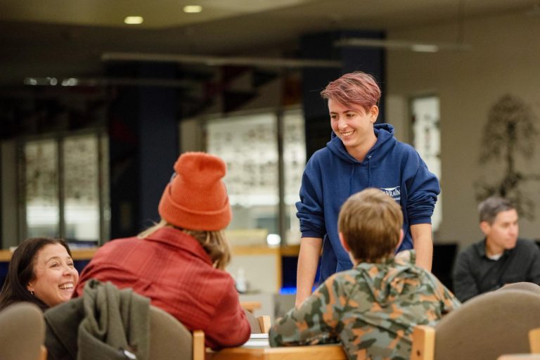 A group of people engaged in conversation at a table, with a smiling person in a blue hoodie standing nearby.