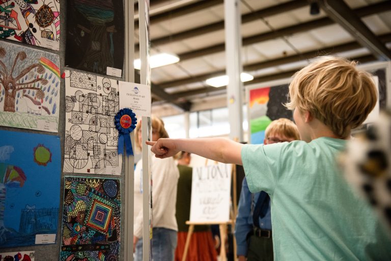 A child points at an award-winning artwork displayed at an art exhibition, surrounded by other attendees.