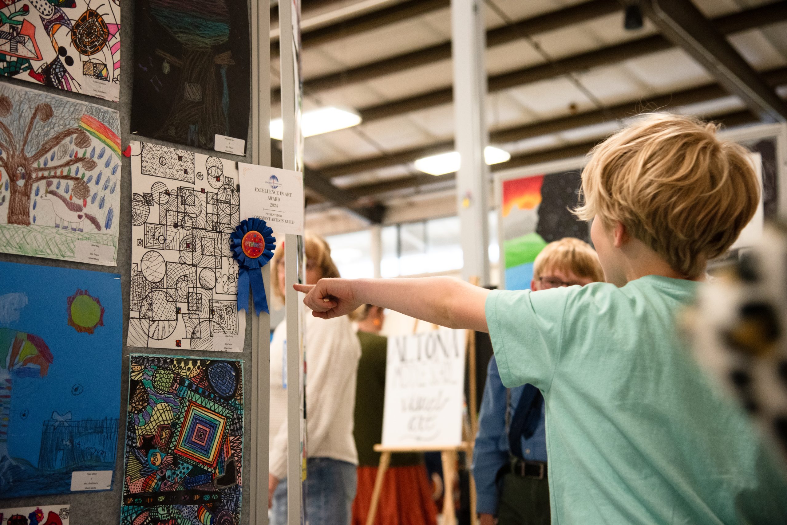 A child points at an award-winning artwork displayed at an art exhibition, surrounded by other attendees.