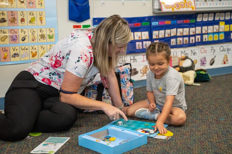 A teacher and a young girl play a colorful board game on the floor in a classroom filled with educational posters.