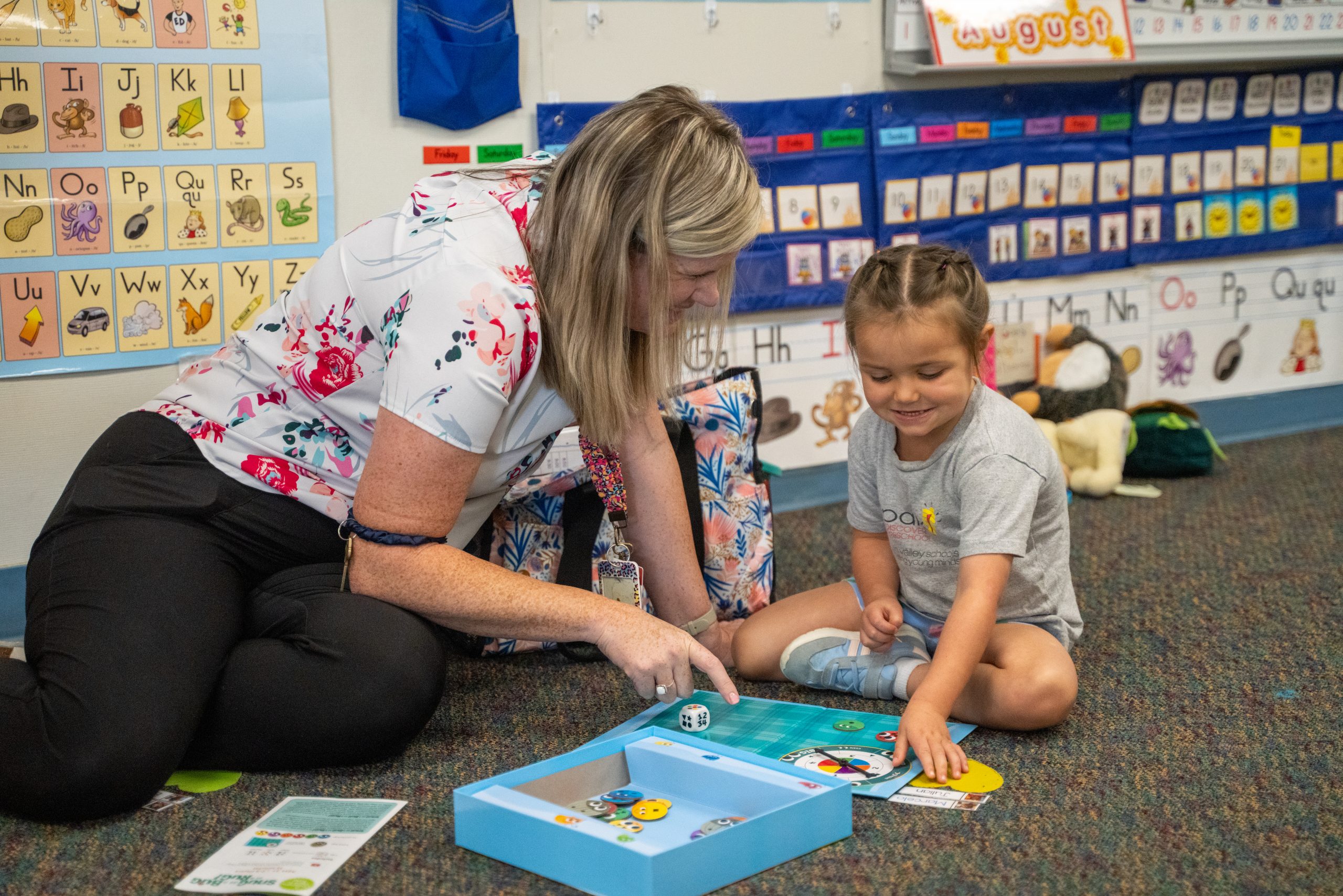 A teacher and a young girl play a colorful board game on the floor in a classroom filled with educational posters.