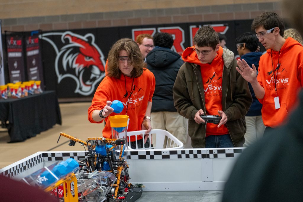 Three students in orange hoodies interact with a robotics competition setup, focusing on controlling a robot.