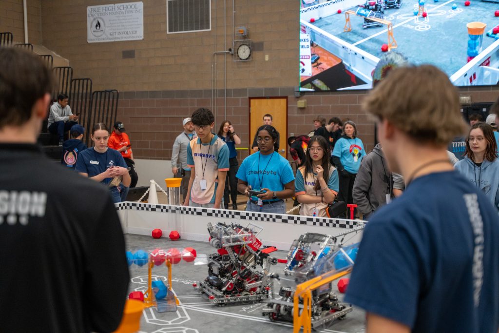 Students observe a robotics competition, with robots on a field and a large screen displaying the event.