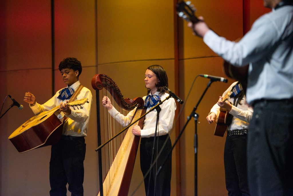 A group of young musicians performs on stage, featuring a guitarist and a harpist, with colorful lighting behind them.
