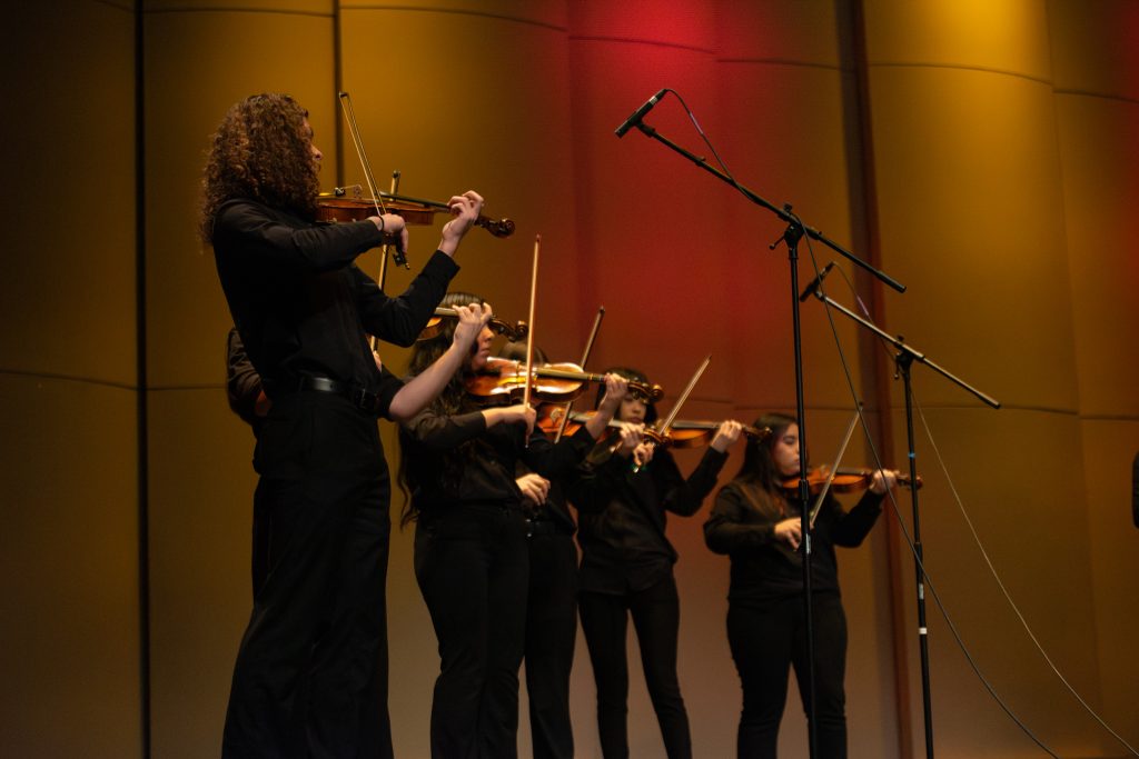 A group of violinists in black attire performs on stage, with colorful lighting in the background.