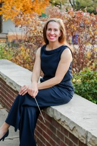 A woman in a navy dress sits on a stone wall amid autumn foliage, holding a conductor's baton and smiling.