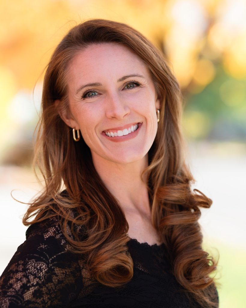 A woman with long, wavy hair smiles warmly, wearing a black lace top, with a blurred autumn background.