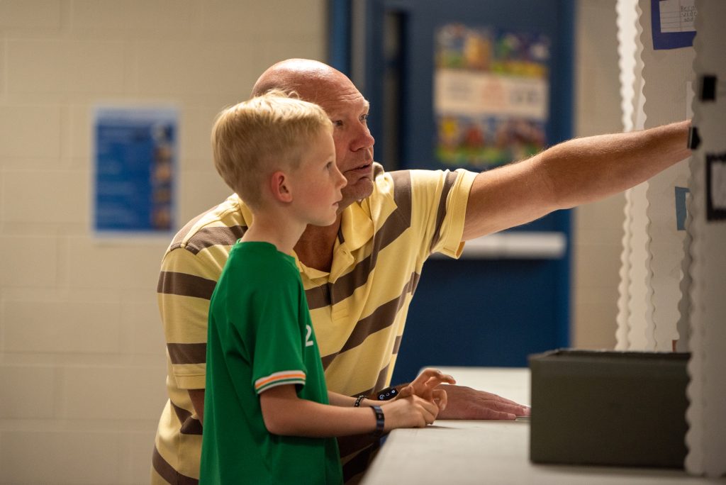 A man and a boy observe a poster board, engaged in conversation, in a well-lit indoor setting.