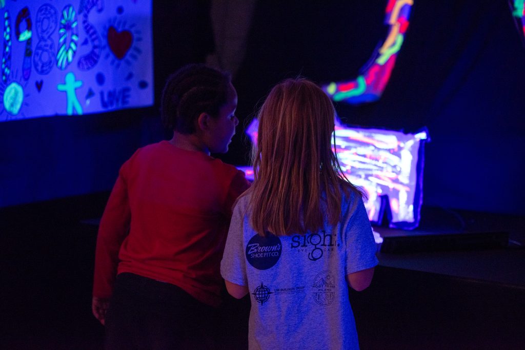 Two children observe colorful artwork illuminated under black light, surrounded by vibrant decorations.