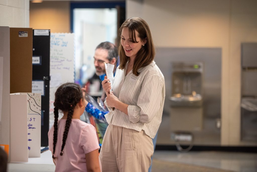 A woman talks to a girl at an event, with displays and people in the background. The setting is indoors.
