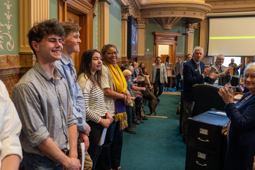 A group of young people stands smiling in a formal setting, while an audience claps in the background.