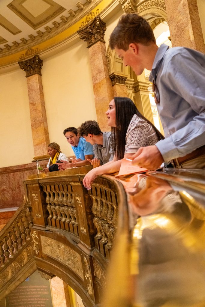 Five young adults gather on a balcony, engaging in conversation in an ornate building with marble columns.