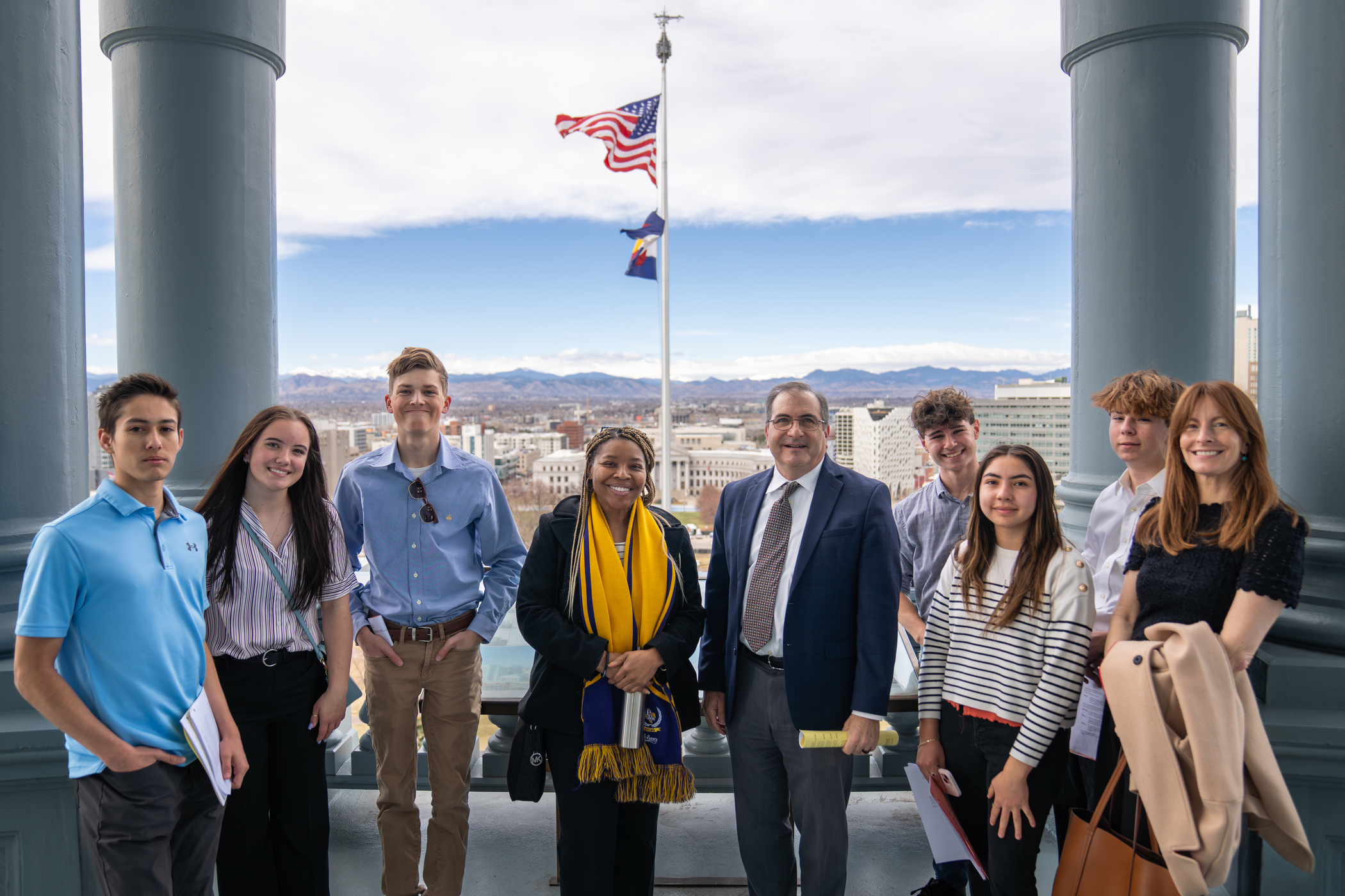 A group of eight people poses outdoors with city and mountains in the background, featuring the American and Colorado flags.
