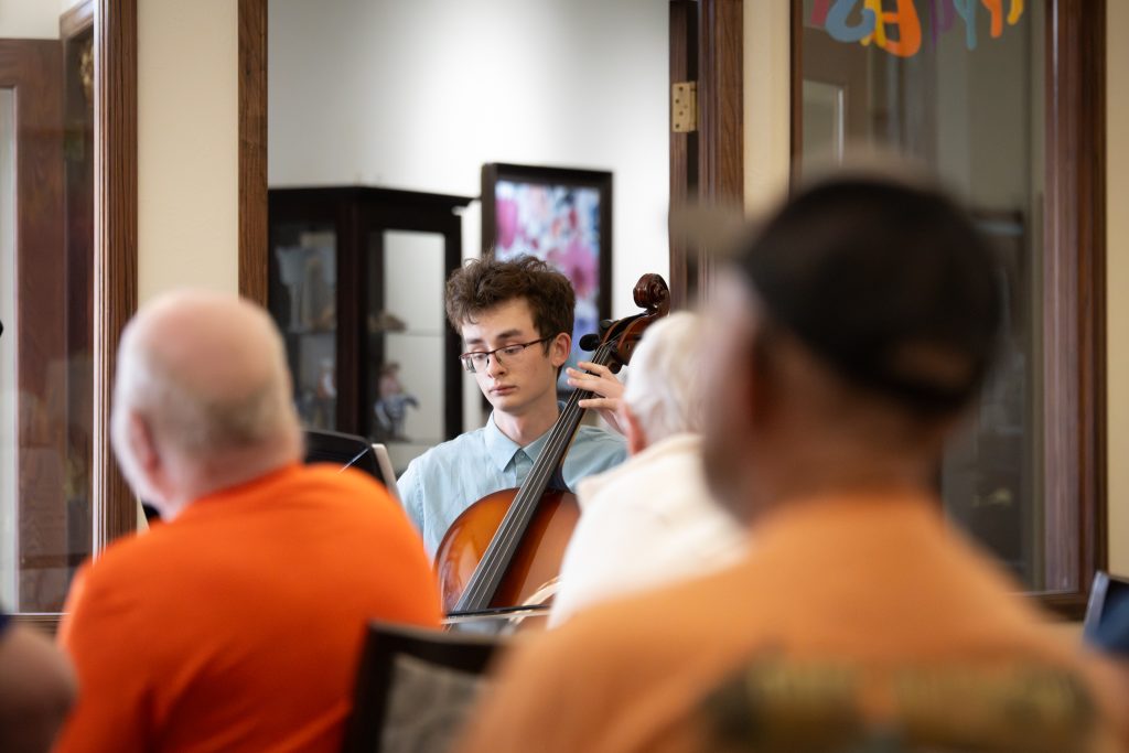 A young man plays the cello in a room filled with seated listeners, some blurred in the foreground.