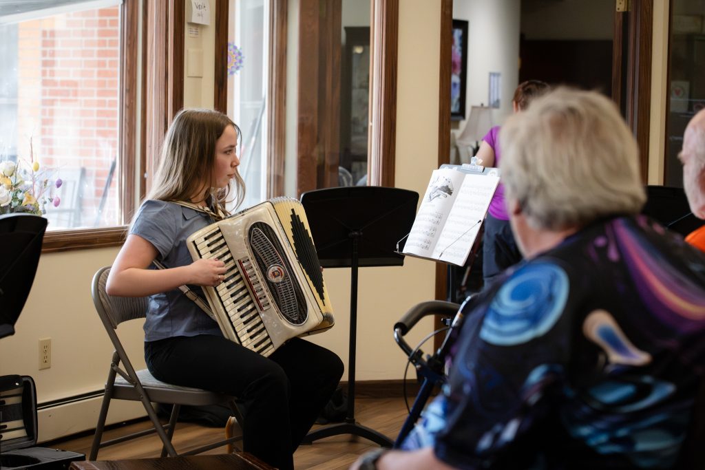 A young woman plays the accordion while seated, with sheet music in front of her and audience members listening.