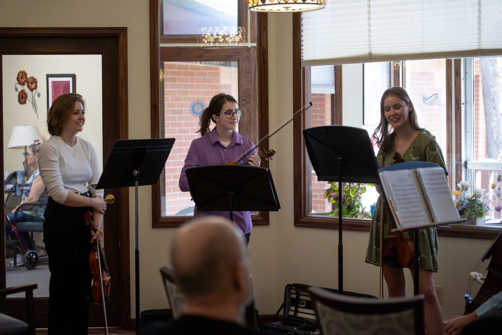 Three musicians perform with violins in a bright room, while audience members enjoy the music.