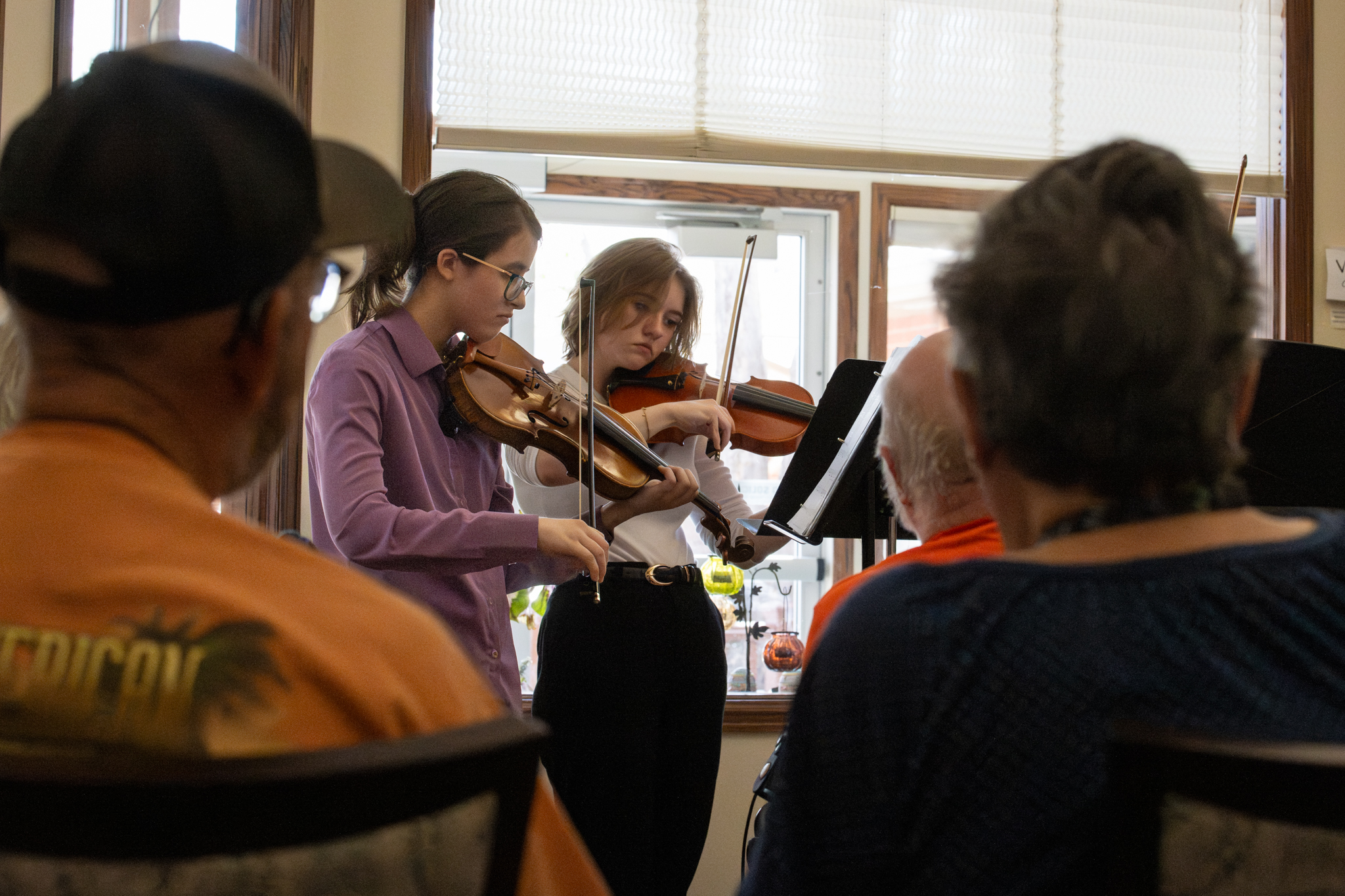 Two young musicians play violins in front of an audience in a cozy indoor setting.