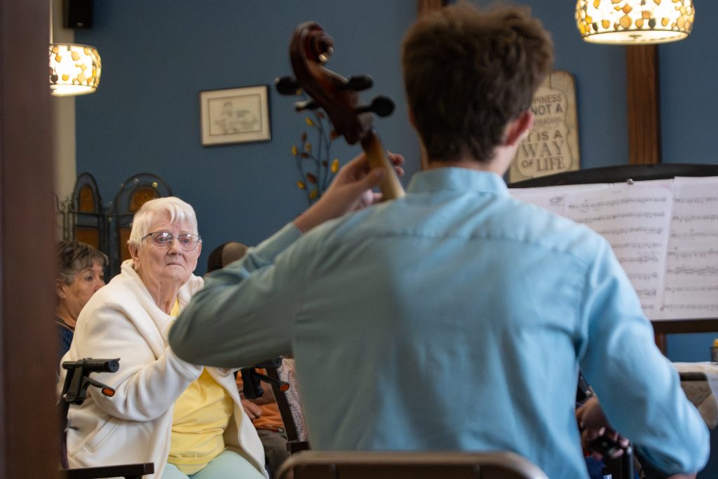 A young musician plays a cello for elderly audience members in a cozy room, with music sheets visible.
