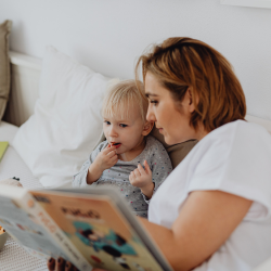 A woman and a toddler sit together on a couch, reading a book while the child eats a snack.