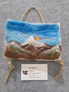 A needle-felted landscape artwork with mountains and a sun, displayed with an informational label.