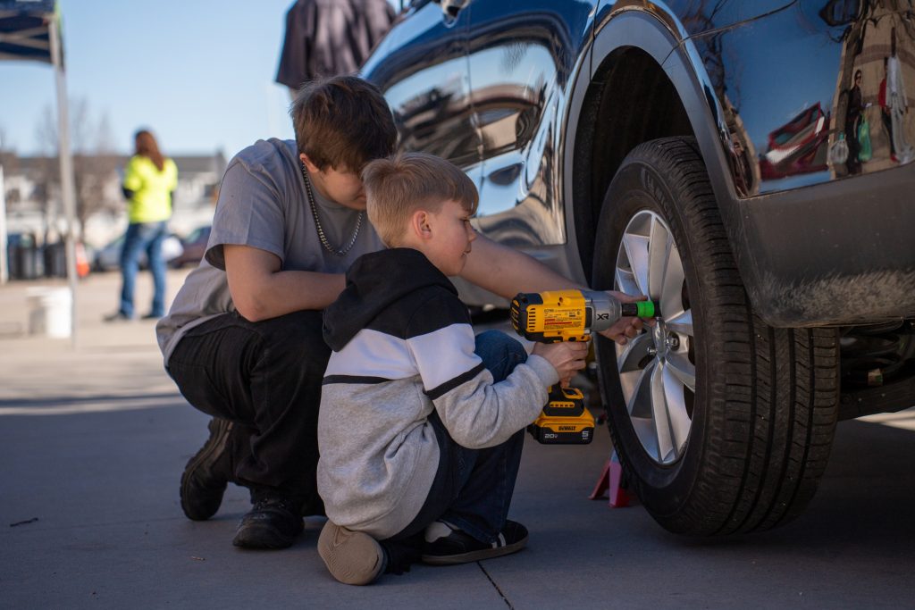 A young boy uses a power drill to work on a car tire while an older teen guides him.