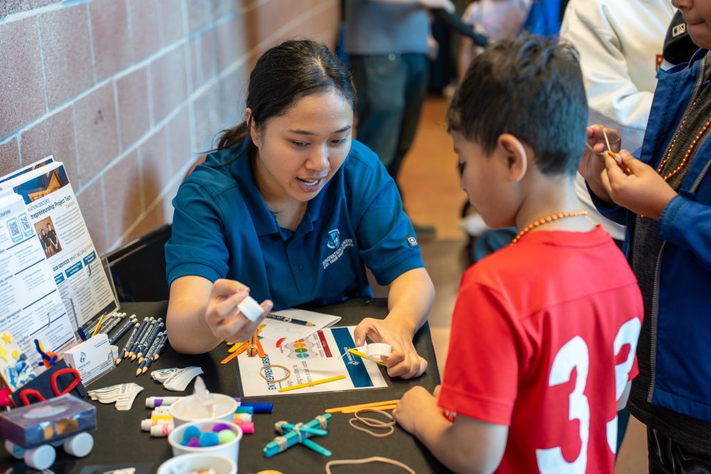 A young woman in a blue shirt explains an activity to a child in a red shirt at a craft table.