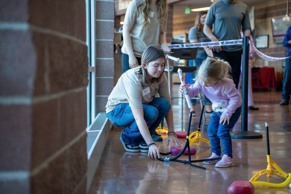 A young girl and an older girl engage with a science activity involving colorful equipment in a school setting.
