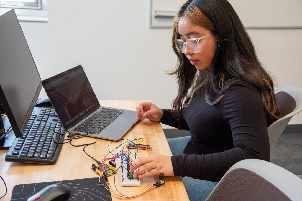A student works on a laptop, assembling wires and components on a breadboard at a wooden desk.