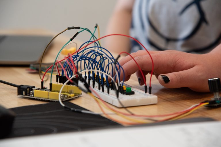 A hand connects wires on a breadboard with a microcontroller and various components on a wooden table, playing a musical instrument prototype.